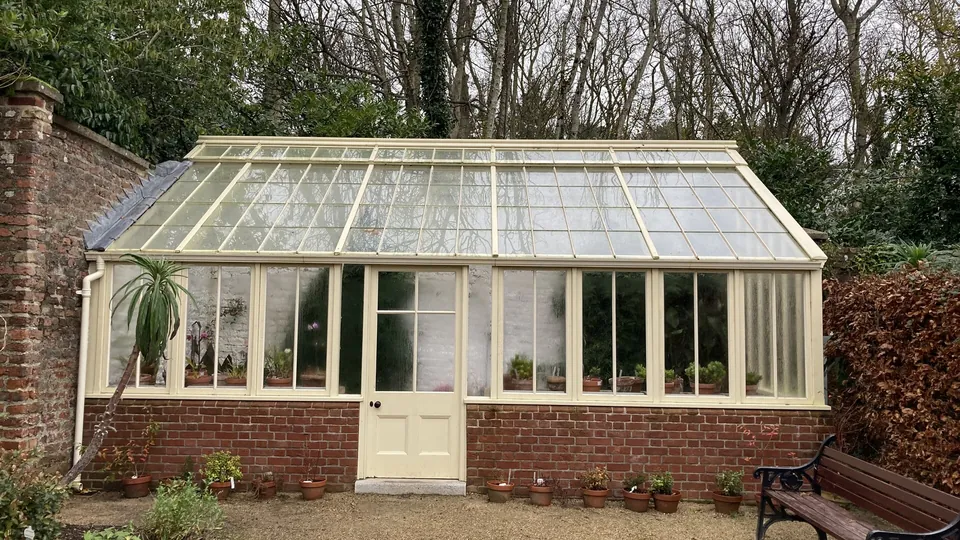Frei stehendes Glasgewächshaus mit Satteldach auf gemauertem Sockel in einem ummauerten Garten, dahinter kahle Bäume